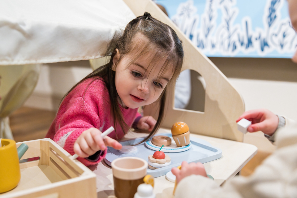 A young girl with brown hair, wearing a pink sweater, plays with toy food and dishes at a small table inside a playhouse.